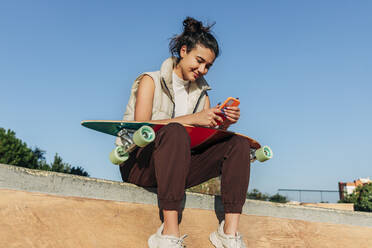 Frau mit Skateboard und Mobiltelefon auf einer Sportrampe im Park - JRVF02753