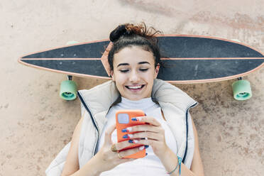 Lächelnde Frau, die auf einem Skateboard liegt und ein Smartphone im Park benutzt - JRVF02755