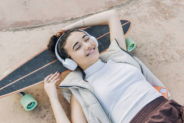 Smiling woman with headphones relaxing on skateboard in park - JRVF02758