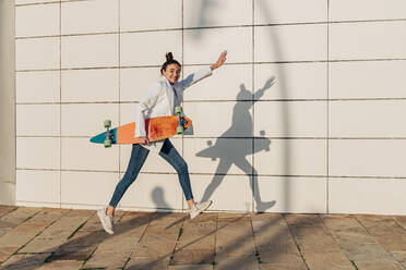 Fröhliche Frau springt mit Skateboard auf Gehweg - JRVF02765