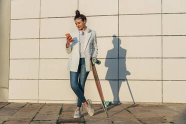 Woman with skateboard using smart phone on footpath - JRVF02773