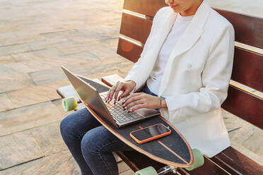 Frau mit Smartphone und Laptop auf dem Skateboard im Park - JRVF02778