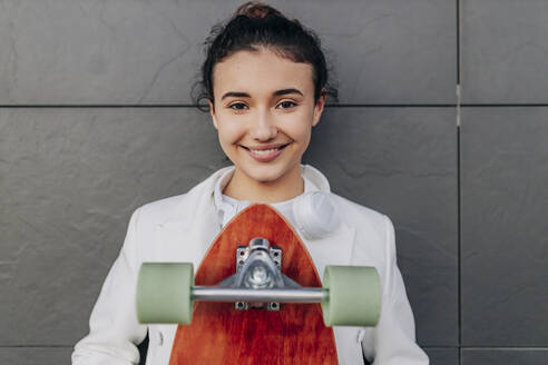 Lächelnde Frau mit Skateboard vor einer schwarzen Wand - JRVF02787