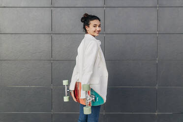 Lächelnde Frau mit Skateboard an der Wand stehend - JRVF02795