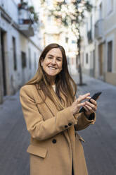 Lächelnde Frau mit Smartphone in Gasse stehend - XLGF02642