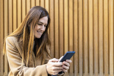 Lächelnde Frau, die mit ihrem Smartphone vor einer Wand Textnachrichten verschickt - XLGF02645