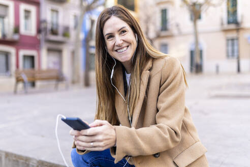 Lächelnde Frau, die über In-Ear-Kopfhörer auf einer Straße in der Stadt mit einem Mobiltelefon spricht - XLGF02652