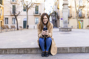 Lächelnde Frau, die auf dem Gehweg sitzend mit ihrem Mobiltelefon im Internet surft - XLGF02654
