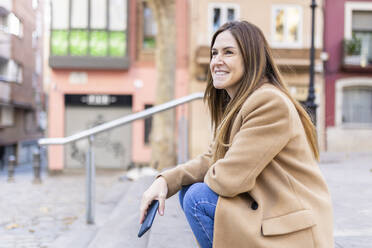 Smiling woman with smart phone sitting on footpath in city - XLGF02657