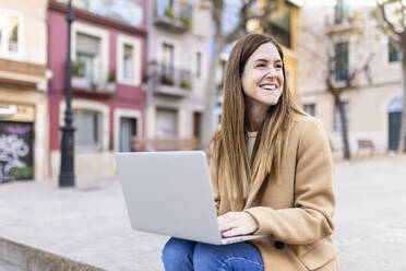Glückliche Frau mit Laptop auf dem Fußweg in der Stadt sitzend - XLGF02660