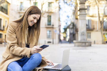 Lächelnde Frau mit Laptop, die über ihr Handy im Internet surft, auf einer Straße in der Stadt - XLGF02662