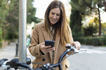 Smiling woman with bicycle using smart phone - XLGF02687