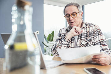 Senior man with hand on chin reading financial bill sitting at table - UUF25305