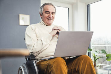 Smiling senior disabled man sitting with laptop on wheelchair at home - UUF25322