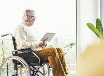 Älterer behinderter Mann mit drahtlosen Kopfhörern, der zu Hause im Rollstuhl einen Tablet-PC benutzt - UUF25336
