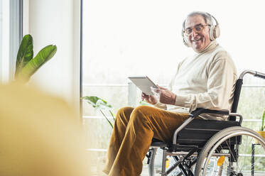 Lächelnder älterer behinderter Mann mit Tablet-PC, der zu Hause im Rollstuhl sitzt - UUF25338