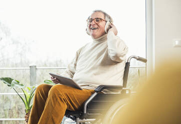 Glücklicher behinderter Mann mit Tablet-PC, der zu Hause im Rollstuhl Musik über kabellose Kopfhörer hört - UUF25341