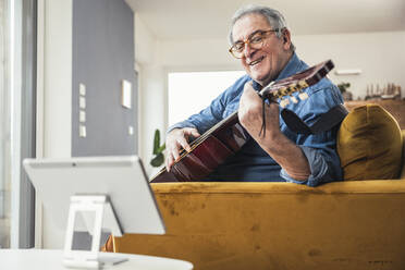 Lächelnder älterer Mann spielt Gitarre und lernt über einen Tablet-PC im Wohnzimmer - UUF25343