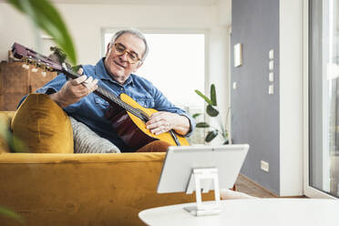 Senior man playing guitar sitting on sofa in living room at home - UUF25344