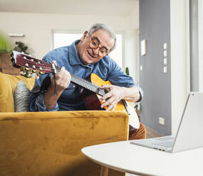 Senior man wearing eyeglasses playing guitar on sofa in living room - UUF25347