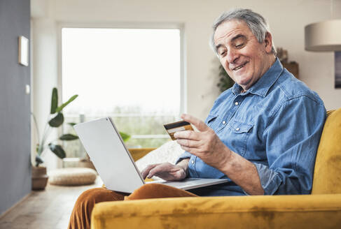 Smiling senior man with credit card and laptop sitting on sofa in living room - UUF25349