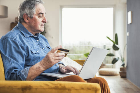 Älterer Mann mit Kreditkarte bei der Online-Zahlung über einen Laptop im Wohnzimmer - UUF25351