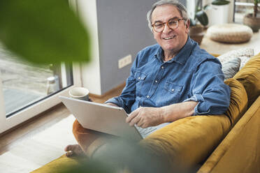 Smiling senior man with laptop and coffee mug sitting on sofa at home - UUF25357