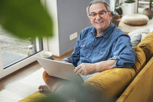 Lächelnder älterer Mann mit Laptop und Kaffeetasse auf dem Sofa zu Hause - UUF25357