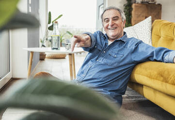 Glücklicher älterer Mann gestikuliert am Sofa im Wohnzimmer zu Hause - UUF25369
