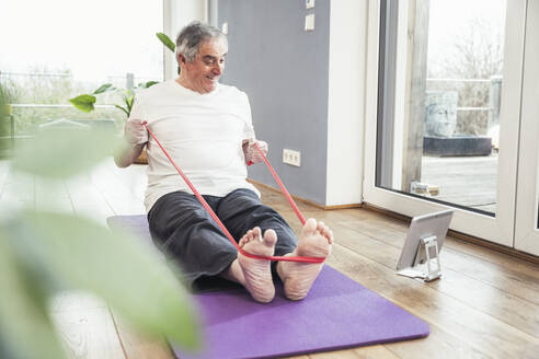 Senior man pulling resistance band watching online tutorial through tablet PC at home - UUF25373