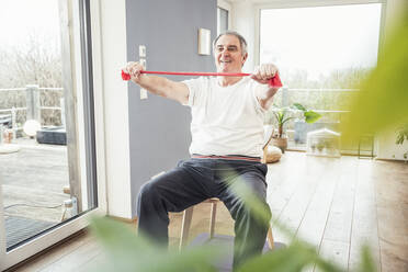 Lächelnder älterer Mann mit Widerstandsband, der auf einem Stuhl sitzt und zu Hause trainiert - UUF25388