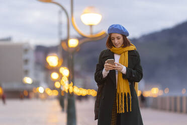 Frau mit blauer Baskenmütze, die in der Abenddämmerung ein Mobiltelefon benutzt - MTBF01142