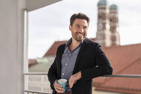 Smiling businessman with coffee cup leaning on railing at office balcony - DIGF17456