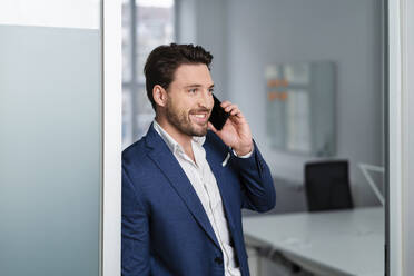 Smiling businessman talking on smart phone at office doorway - DIGF17483