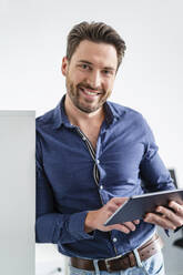 Lächelnder Geschäftsmann mit Tablet-PC, der sich auf einen Schrank im Büro stützt - DIGF17501