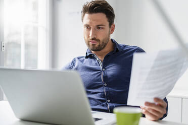 Businessman with document working on laptop in office - DIGF17524