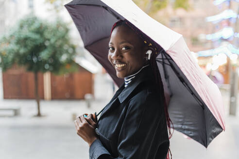 Frau mit drahtlosen In-Ear-Kopfhörern hält Regenschirm - JRVF02806