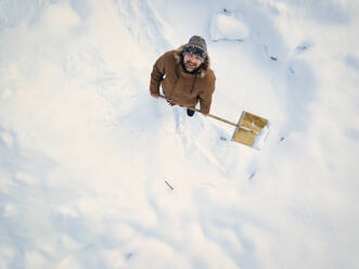 Lächelnder Mann, der eine Schneeschaufel hält und im Winter auf Schnee steht - KNTF06601