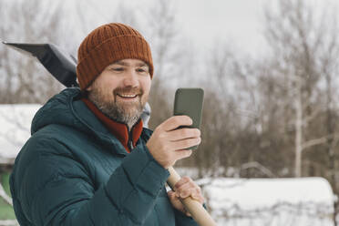 Happy man carrying snow shovel using smart phone in winter - KNTF06614