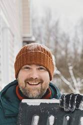 Happy man wearing knit hat holding snow shovel in winter - KNTF06617