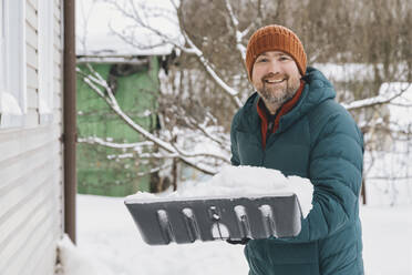 Lächelnder Mann hält Schneeschaufel mit Schnee im Winter - KNTF06618