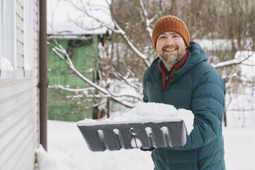 Smiling man holding snow shovel with snow in winter - KNTF06618