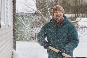 Mann mit Schneeschaufel im verschneiten Garten stehend - KNTF06619