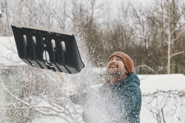 Playful man throwing snow from snow shovel in winter - KNTF06620