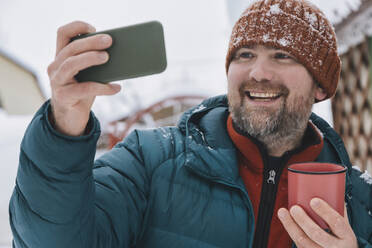 Lächelnder Mann, der einen Kaffeebecher hält und ein Selfie mit seinem Mobiltelefon im Winter macht - KNTF06624