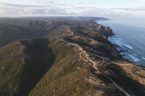 Portugal, Algarve, Vila do Bispo, Aerial view of dirt road stretching across coastal hills surrounding Praia do Barranco beach at dawn - MKLF00044