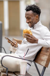 Lächelnder Mann mit Burger und Mobiltelefon auf einer Bank sitzend - OCMF02324