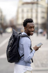 Lächelnder junger Mann mit Rucksack und Mobiltelefon - OCMF02327