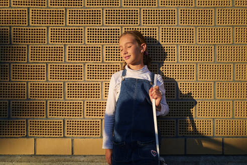 Girl with eyes closed standing in front of brick wall at sunset - DIKF00618