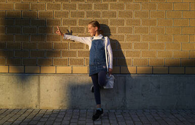 Glückliches Mädchen, das auf einem Bein stehend vor einer Backsteinmauer ein Friedenszeichen macht - DIKF00620